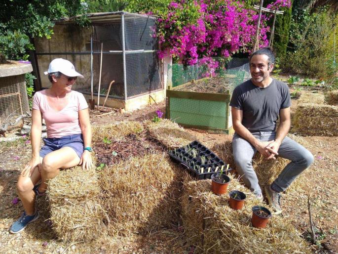 Heather and Joe sitting on one of the straw beds in the kitchen garden with the first of the Hugelkultur beds in the background